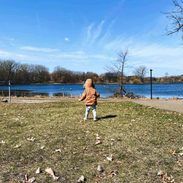A young child in a brown jacket with the hood over their head walks near the water alone.