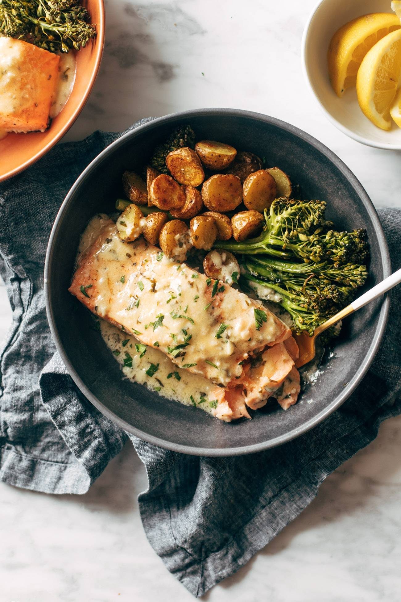 Baked salmon in a bowl with broccoli, roasted potatoes, and lemon sauce on top. There's a fork in the bowl with a bowl of lemons on the side.